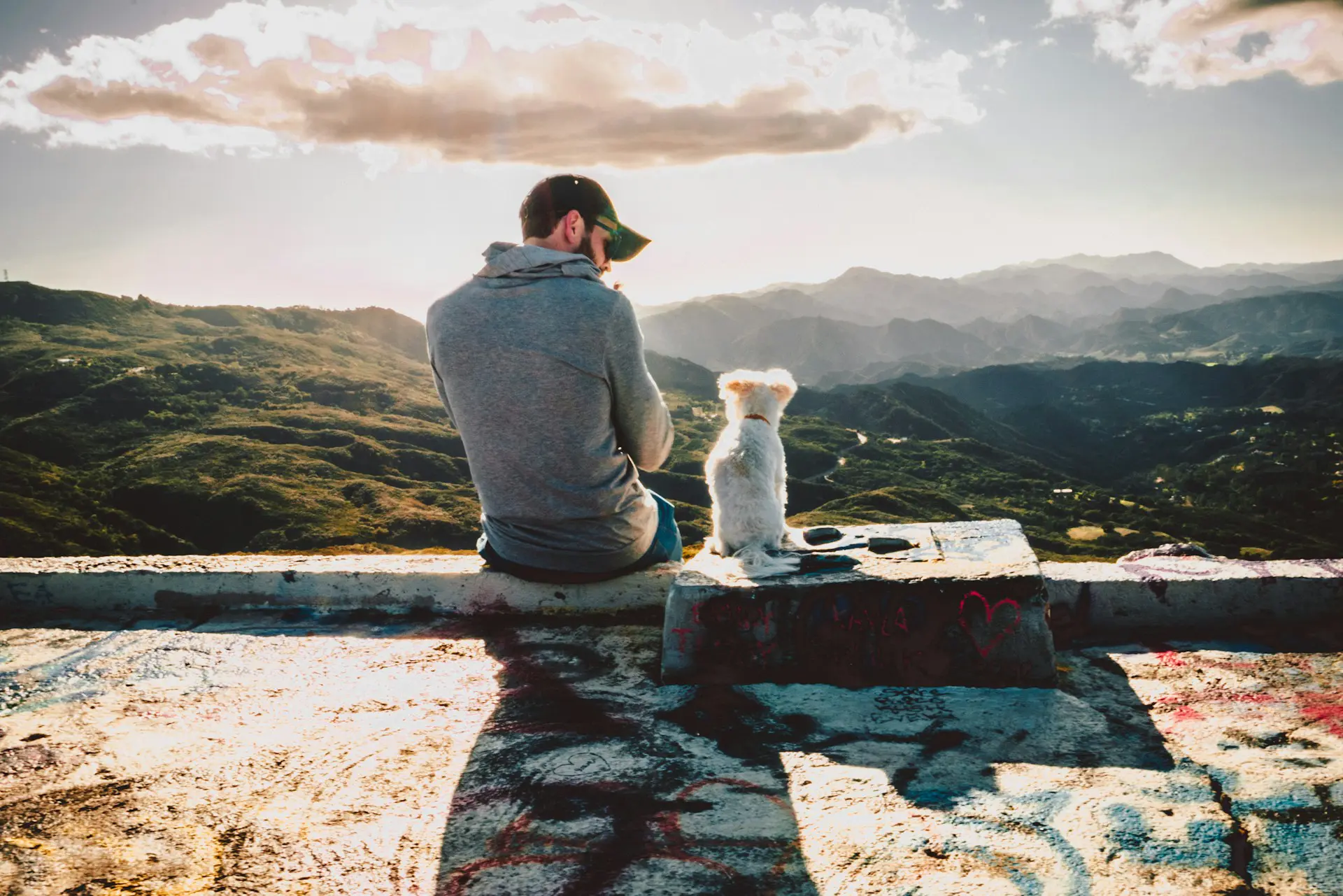 man wearing hat sitting with dog on ledge overlooking mountains